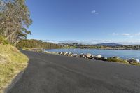 the side view of a paved road next to water with rocks around it and some trees in front of a large body of water