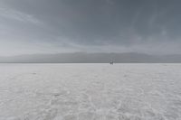 two people are standing on the salt with mountains in the background while one person is walking across the expanse