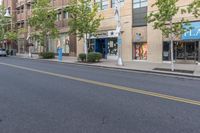 a street with trees and a building on the other side of it, and a blue traffic light hanging off the pole above