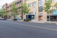 a street with trees and a building on the other side of it, and a blue traffic light hanging off the pole above