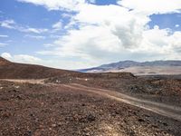 an open dirt road stretches across a desert area with rolling hills and boulders and a cloud covered sky