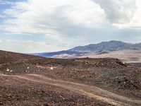 an open dirt road stretches across a desert area with rolling hills and boulders and a cloud covered sky