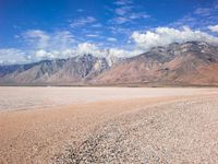 a desert is seen under cloudy skies with mountains in the background and blue skies, with clouds, sand and stones