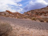 a desert with rocks and grass and shrubs under a cloudy sky over them is shown