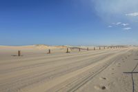 sand dunes and wooden poles with shadows in it, along side of a beach, under a blue sky with cloud