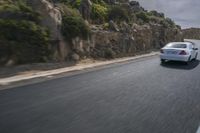 a white car driving down the road near rocks and mountains in the background, some trees, rocks, and bushes on both sides