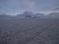 a picture of a field of desert with dark clouds and mountains in the distance, including a mountain, an area, dirt, and a few tracks, in the ground