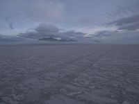 a picture of a field of desert with dark clouds and mountains in the distance, including a mountain, an area, dirt, and a few tracks, in the ground