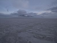 a picture of a field of desert with dark clouds and mountains in the distance, including a mountain, an area, dirt, and a few tracks, in the ground