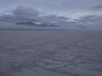 a picture of a field of desert with dark clouds and mountains in the distance, including a mountain, an area, dirt, and a few tracks, in the ground