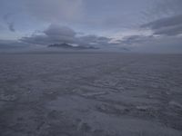 a picture of a field of desert with dark clouds and mountains in the distance, including a mountain, an area, dirt, and a few tracks, in the ground