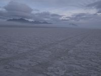 a picture of a field of desert with dark clouds and mountains in the distance, including a mountain, an area, dirt, and a few tracks, in the ground