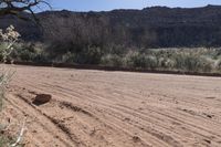 an empty dirt road in front of a mountain with a large brown piece of rock lying on the ground