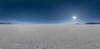 a sun shines over a large sandy flat plain under a cloudy blue sky behind