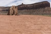 an odd rock on a desert area with mountains in the background, with white clouds