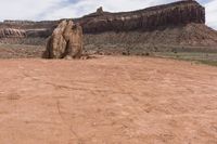 an odd rock on a desert area with mountains in the background, with white clouds