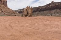 an odd rock on a desert area with mountains in the background, with white clouds