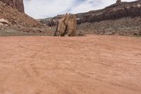 an odd rock on a desert area with mountains in the background, with white clouds