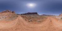 a picture of dirt road going through desert land with rock cliff in background and sun shining