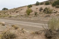 a dirt road through the desert with rocks and grass on it's sides and hills in the background