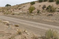 a dirt road through the desert with rocks and grass on it's sides and hills in the background