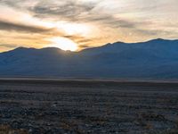 a white and black horse grazing in the desert with a sunset behind it and mountains