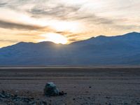 a white and black horse grazing in the desert with a sunset behind it and mountains