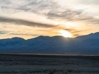 a white and black horse grazing in the desert with a sunset behind it and mountains