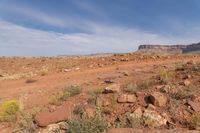 an open dirt field near a rocky cliff in the desert below blue skies, with a small mountain in the distance