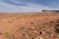 an open dirt field near a rocky cliff in the desert below blue skies, with a small mountain in the distance