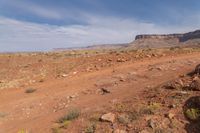 an open dirt field near a rocky cliff in the desert below blue skies, with a small mountain in the distance