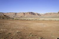 a dirt field with hills and plants in the background, near a blue sky,