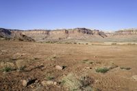 a dirt field with hills and plants in the background, near a blue sky,