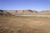 a dirt field with hills and plants in the background, near a blue sky,