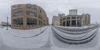 a snow covered street with a big building in the background as well as snow falling on it