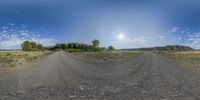 the view of a large dirt road, with grass, bushes and a sun in the sky