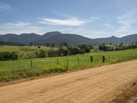 dirt road through field with mountains and sky in distance in background with person walking down it