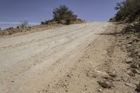 dirt road through the desert with brush and trees in background along side of hill with dirt, vegetation and sky