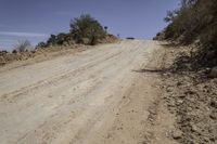 dirt road through the desert with brush and trees in background along side of hill with dirt, vegetation and sky