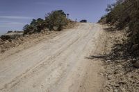 dirt road through the desert with brush and trees in background along side of hill with dirt, vegetation and sky