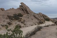 a dirt road and some rocks in the desert on a cloudy day and there is a person on a motorcycle