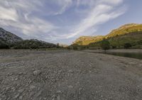 a dirt field with trees and rocks near a body of water and mountains in the distance