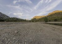 a dirt field with trees and rocks near a body of water and mountains in the distance