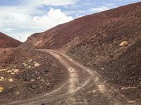 a dirt road on the side of a hill in front of some mountains and a sky