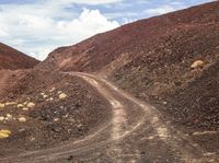 a dirt road on the side of a hill in front of some mountains and a sky