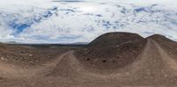 an outdoor panorama of dirt road and mountains with trees in the distance against a cloudy sky