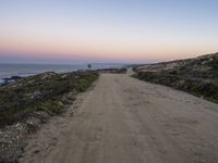 a dirt road leading towards a ocean by the shoreline at sunset in california on a hazy day