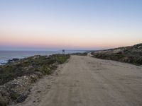 a dirt road leading towards a ocean by the shoreline at sunset in california on a hazy day