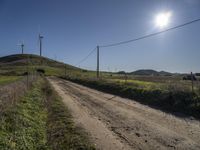 dirt road near a fence with wind turbines in the distance with blue skies and sun