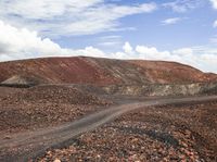 a dirt and rock road with trees in the distance near a hill with mountains on one side